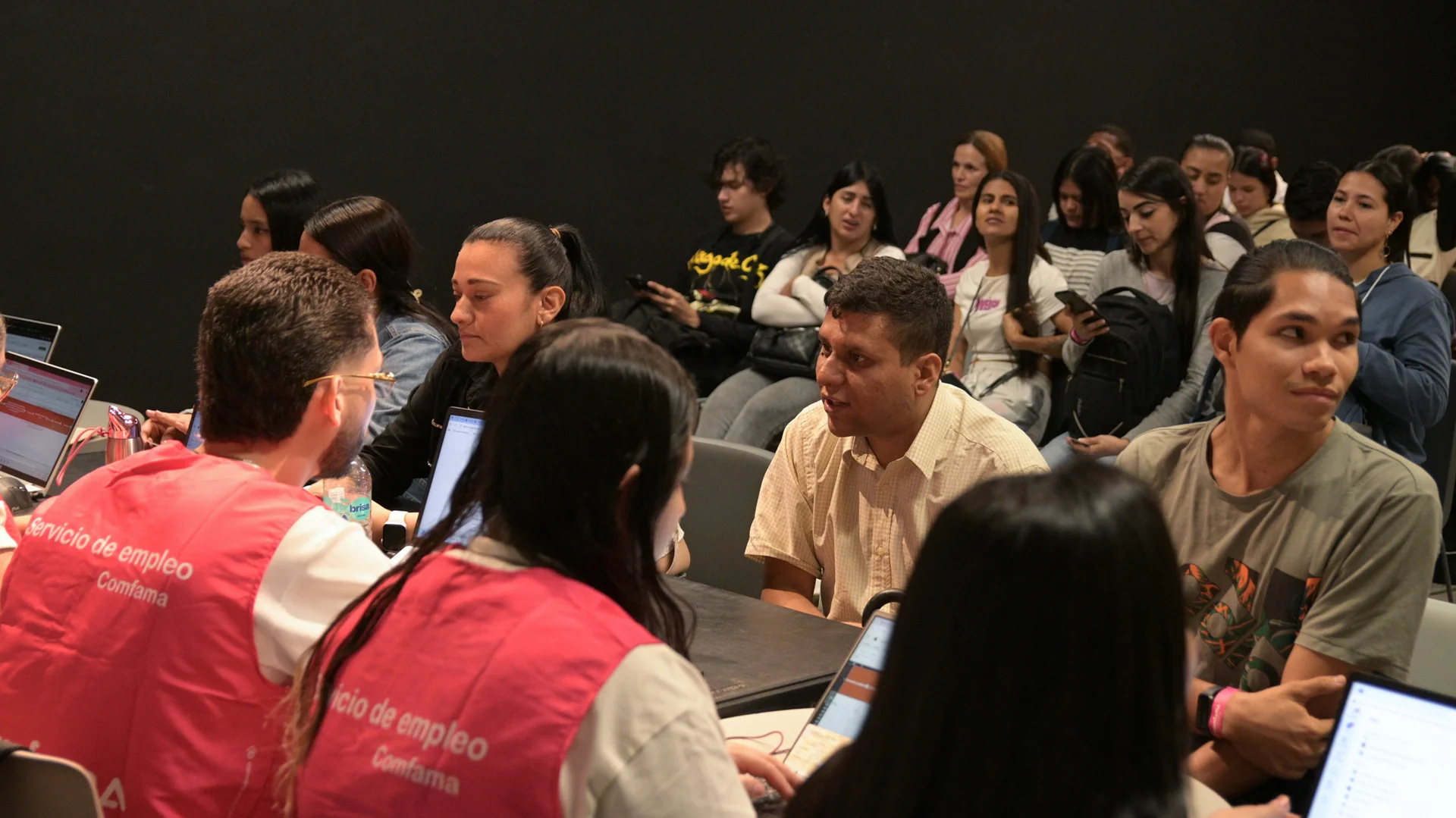 Personas participan en una actividad grupal en un auditorio, mientras conversan y trabajan con computadores portátiles