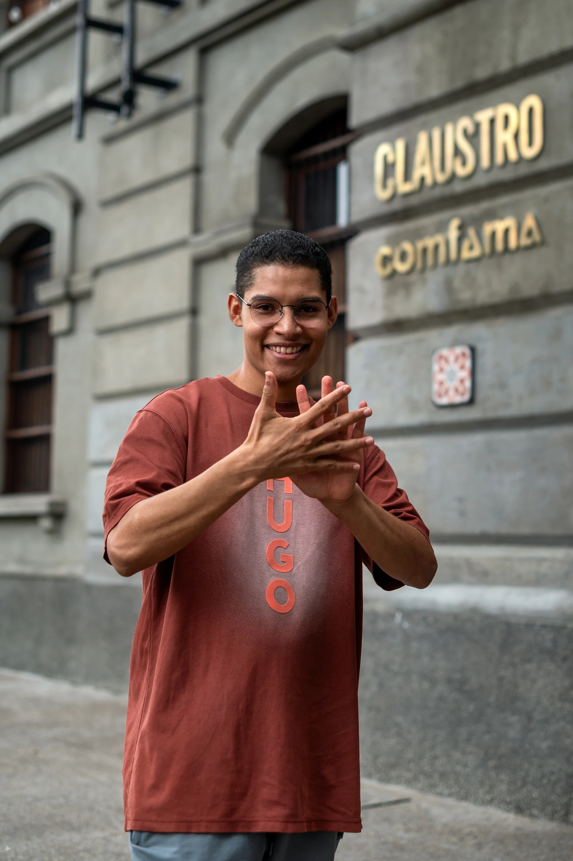 Joven sonriente frente a la entrada del claustro Comfama, realizando lenguaje de señas